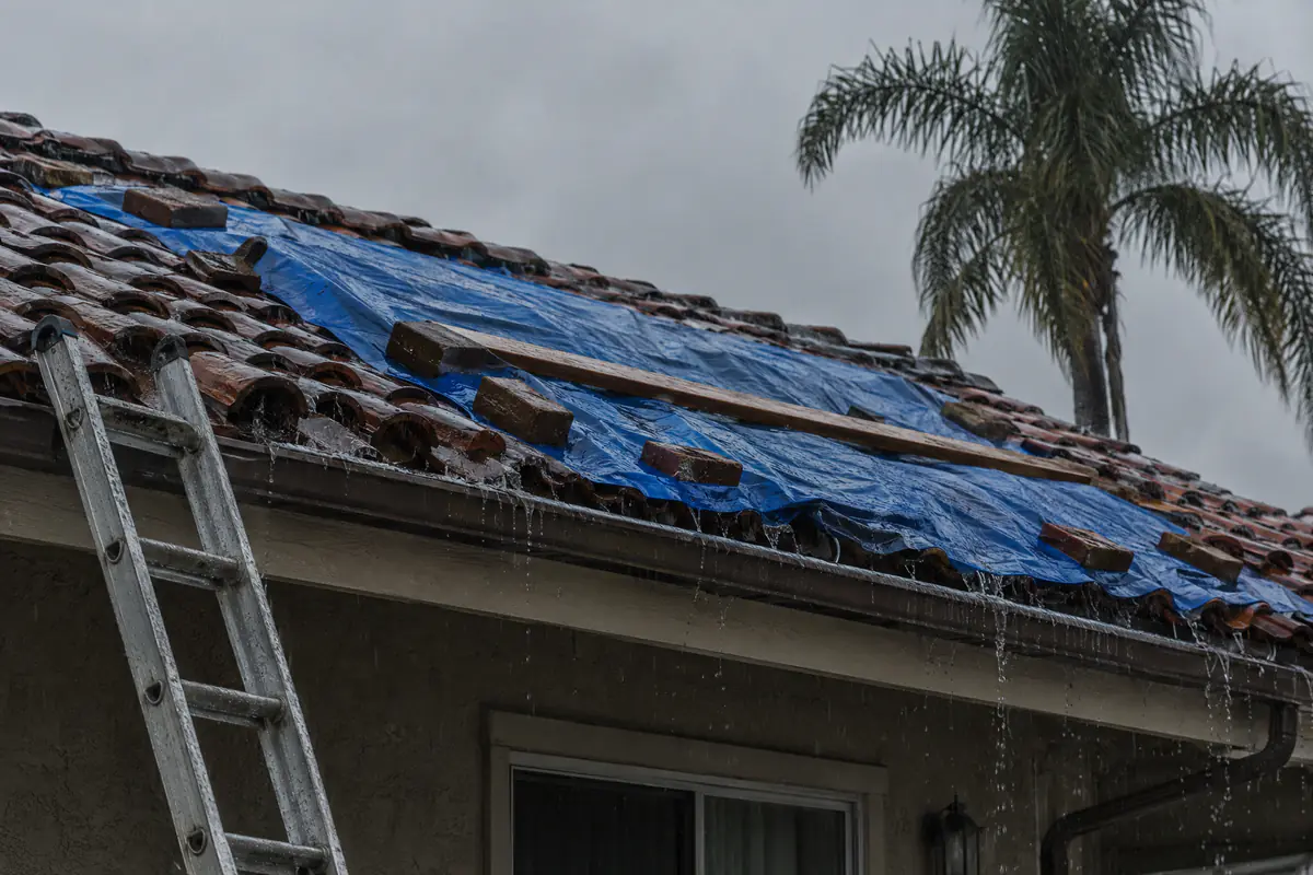 Rain on a Spanish tile roof with a blue tarp weighted by bricks and a ladder at the eave.