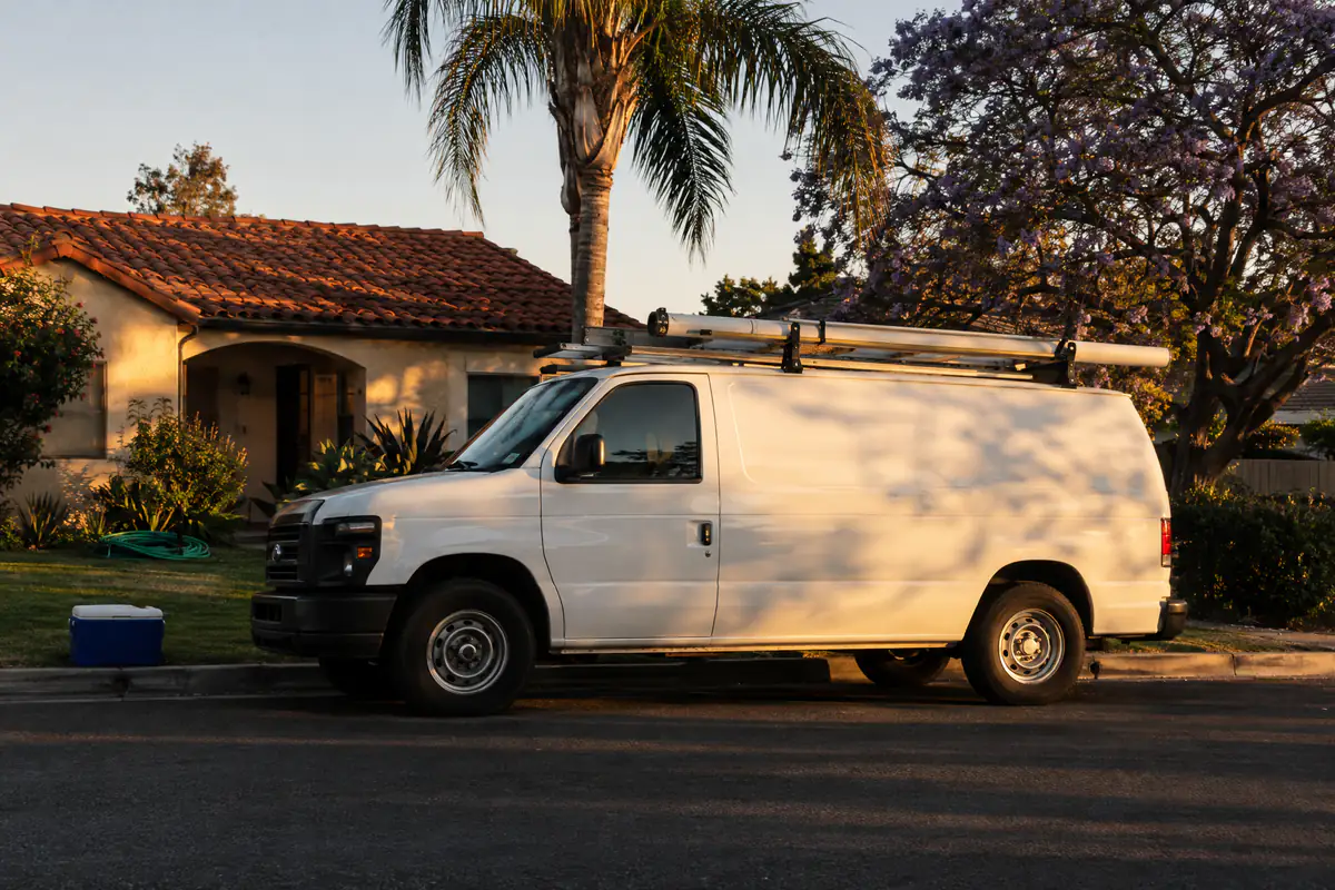A blank white work van with a ladder rack parked outside a Spanish-tile bungalow in San Diego at golden hour.